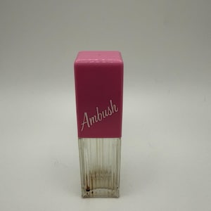 Puede incluir: Un frasco de perfume rectangular con una base de vidrio transparente con estrías verticales y una tapa rosa sólida. La palabra Ambush está impresa en letra cursiva blanca en la parte frontal de la tapa rosa. El frasco tiene un diseño alto y esbelto.