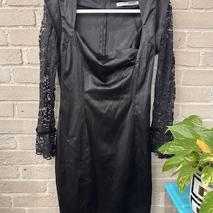 May include: A black satin dress with a square neckline and long sleeves. The sleeves are made of black lace. The dress is hanging on a white hanger against a brick wall.