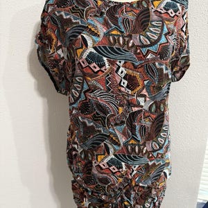 May include: A short-sleeved top with a colorful, intricate pattern. The design features a mix of geometric shapes and organic forms in shades of brown, black, blue, orange, and white. The top has a gathered detail at the waist and the text "Lake Odessa" on the mannequin.