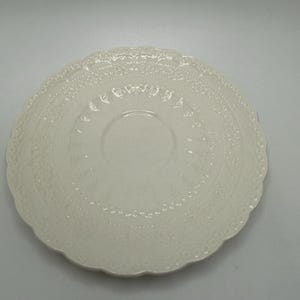 May include: A round, off-white ceramic saucer featuring a scalloped edge and an intricate, embossed lace-like pattern that radiates from the center. The surface has a glossy finish with a recessed circular area designed to hold a matching cup.