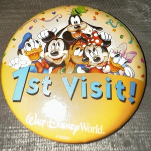 Könnte beinhalten: Ein runder, gelber Button mit dem Text "1st Visit!" und dem Walt Disney World-Logo. Der Button zeigt die Zeichentrickfiguren Mickey Mouse, Minnie Mouse, Donald Duck, Goofy, Pluto und Daisy Duck.