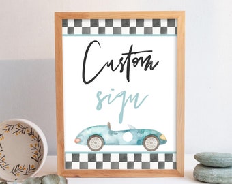 May include: A framed sign with a light wood frame. The sign features the words "Custom sign" in black and light blue script. Below the text is a watercolor illustration of a light blue vintage race car. The top and bottom borders have a black and white checkered pattern.