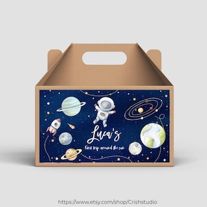 Puede incluir: Una caja de cartón marrón para favores de fiesta con un diseño de astronauta, planetas y estrellas azules y blancas. La caja tiene un asa y dice "Luca's First trip around the sun".