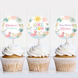 May include: Three cupcakes with white frosting, each topped with a circular topper. The toppers feature tropical designs with flamingos, pineapples, and the text "Madison's First Birthday", "One in the Sun", and "Happy Birthday Madison".