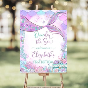 May include: A purple and teal mermaid tail with gold glitter accents is featured on a welcome sign for a first birthday party. The sign reads "Oneder the Sea - welcome to Elizabeth's First Birthday".