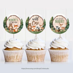 Pode incluir: Tr&ecirc;s toppers redondos para cupcakes com ilustra&ccedil;&otilde;es de animais da floresta e texto. Os toppers apresentam um urso marrom, uma raposa, um guaxinim, um veado e um tronco de &aacute;rvore com o texto "Wild One" e "Robert's 1st Birthday".