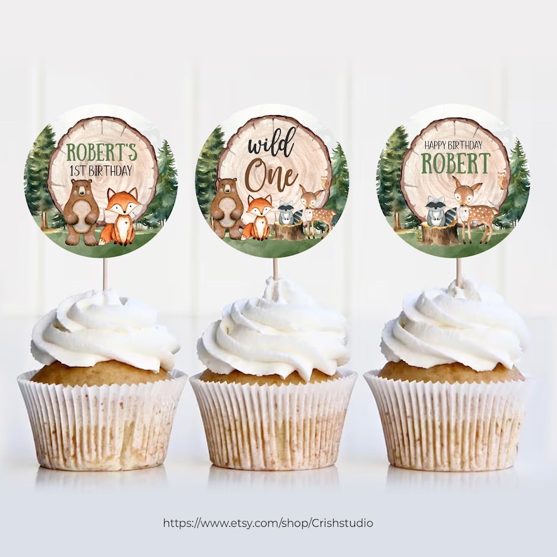 Pode incluir: Tr&ecirc;s toppers redondos para cupcakes com ilustra&ccedil;&otilde;es de animais da floresta e texto. Os toppers apresentam um urso marrom, uma raposa, um guaxinim, um veado e um tronco de &aacute;rvore com o texto "Wild One" e "Robert's 1st Birthday".