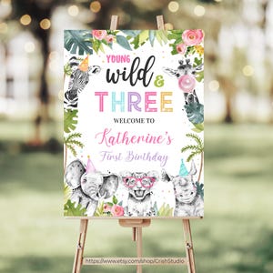 May include: A welcome sign for a first birthday party with a jungle theme. The sign features a white background with green leaves, pink flowers, and a giraffe, zebra, elephant, and rhinoceros. The text on the sign reads "Young Wild & Three, Welcome to Katherine's First Birthday".