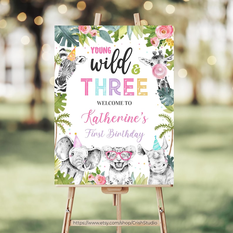 May include: A welcome sign for a first birthday party with a jungle theme. The sign features a white background with green leaves, pink flowers, and a giraffe, zebra, elephant, and rhinoceros. The text on the sign reads "Young Wild & Three, Welcome to Katherine's First Birthday".