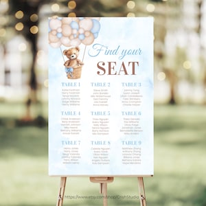 May include: A blue and beige seating chart with a teddy bear in a hot air balloon. The chart lists tables with names for each table. The text "Find your seat" is at the top of the chart.