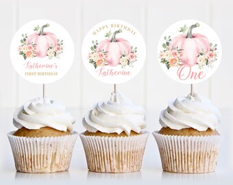 May include: Three cupcakes with white frosting and decorative toppers. The toppers feature a pink pumpkin design with floral accents and text that reads "Katherine's First Birthday", "Happy Birthday Katherine", and "One".