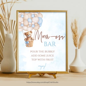 May include: A printable sign for a Mom-osa bar at a baby shower. The sign features a teddy bear in a hot air balloon with blue and beige balloons. The text on the sign reads "Mom-osa Bar", "Pour the bubbly, add some juice, top with fruit", and "enjoy!"