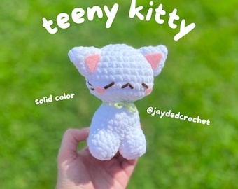 May include: A white crocheted kitty with pink ears and a green bow. The kitty is being held in a hand against a green grassy background. The text "teeny kitty" is written above the kitty and "solid color" is written below the kitty. The text "@jaydedcrochet" is written below the kitty on the right side of the image.