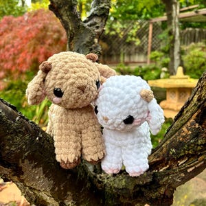 May include: Two crocheted lamb figures, one beige and one white, perched on a tree branch. The lambs have black eyes, small ears, and pink accents on their feet. The background shows a blurred garden with green and red leaves.