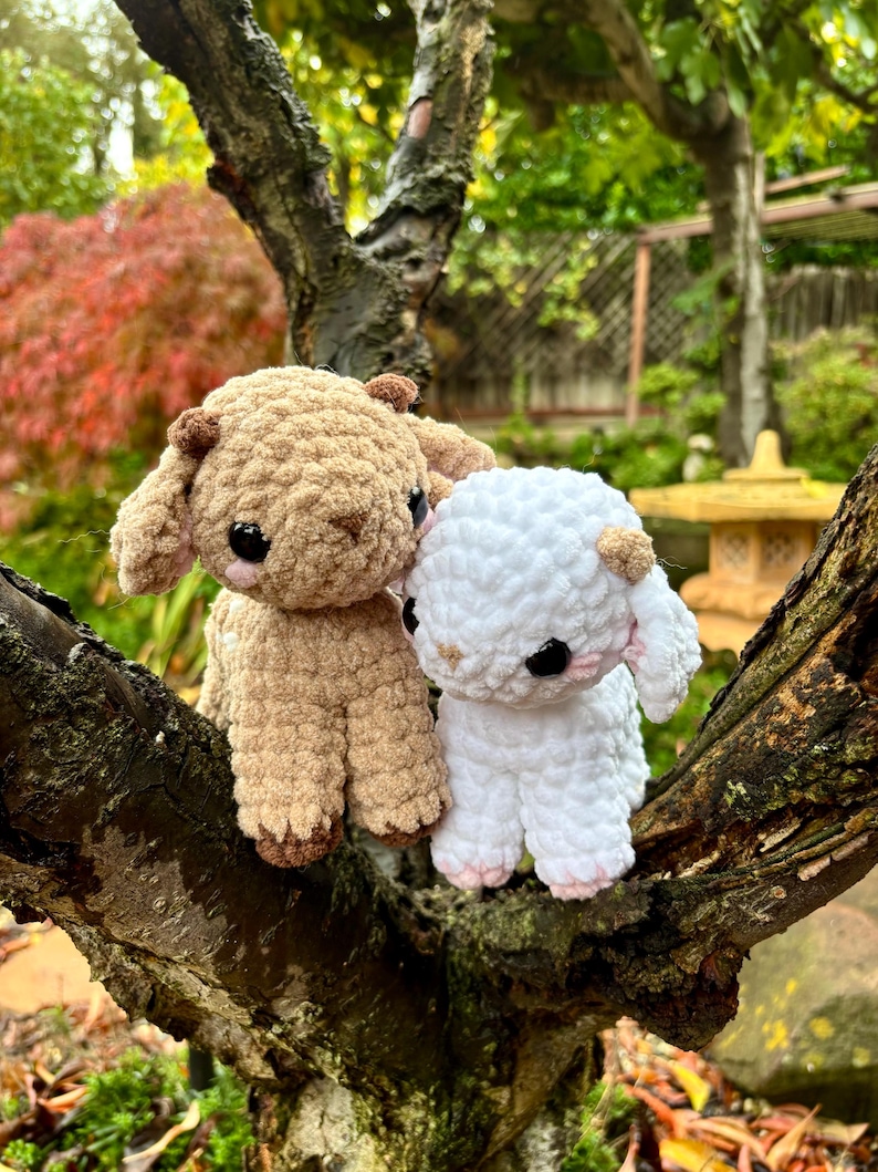 May include: Two handmade crochet lamb figurines, one tan and one white, sit on a tree branch. The lambs have black eyes, small ears, and pink accents on their feet. The background features a blurred garden scene with green and red foliage.