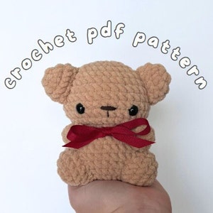 May include: Crocheted teddy bear with brown fur, black eyes, and a red bow. The bear is being held in a hand. The text "crochet pdf pattern" is written in white letters around the bear.