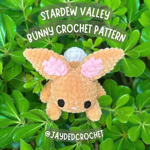 May include: A crocheted brown bunny with pink ears and a white tail. The bunny is sitting on a bed of green leaves. The text "Stardew Valley Bunny Crochet Pattern" and "@jaydedcrochet" are visible in the image.