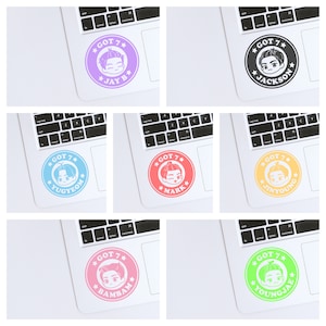 Puede incluir: Un conjunto de nueve pegatinas para portátil con ilustraciones de dibujos animados de los miembros del grupo de K-pop GOT7. Cada pegatina presenta la cara de un miembro diferente en un círculo con el nombre del grupo y el nombre del miembro escrito en texto blanco. Las pegatinas están sobre un fondo blanco.