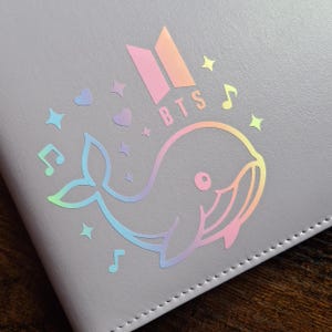 May include: A light gray notebook featuring a holographic whale design with musical notes, stars, and hearts. The word "BTS" is printed in a gradient color scheme. The notebook has a stitched border.
