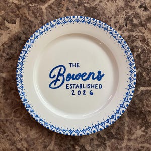 Puede incluir: Plato de cerámica blanco con un borde decorativo azul y el texto "The Bowers Established 2026" en escritura azul. El plato tiene un diseño simple y elegante, adecuado para exhibición o uso.