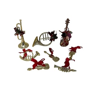 May include: A collection of gold-colored musical instrument ornaments, including a trumpet, French horn, violin, and guitar. Each ornament is adorned with a red ribbon and decorative accents, perfect for holiday decorating. The ornaments are set against a white background.