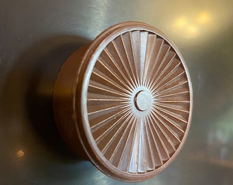 Könnte beinhalten: Ein brauner Kunststoffknopf mit einem Sonnenstrahl-Design. Der Knopf hat eine erhabene Mitte mit einem kleinen Loch in der Mitte.
