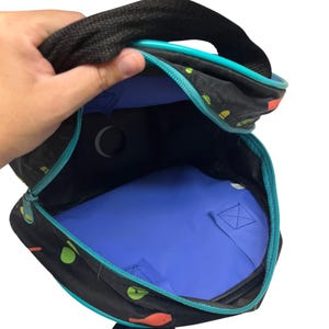 Puede incluir: Una mochila negra con un patr&oacute;n colorido de formas peque&ntilde;as, con un forro interior azul brillante, un borde de cremallera turquesa y correas de tela negra. La bolsa est&aacute; abierta, revelando un recorte circular en el panel trasero y una base azul.