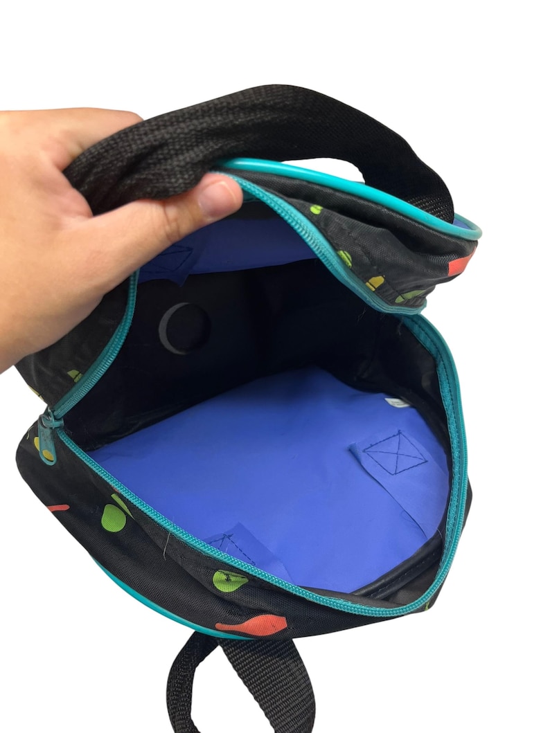 Puede incluir: Una mochila negra con un patr&oacute;n colorido de formas peque&ntilde;as, con un forro interior azul brillante, un borde de cremallera turquesa y correas de tela negra. La bolsa est&aacute; abierta, revelando un recorte circular en el panel trasero y una base azul.