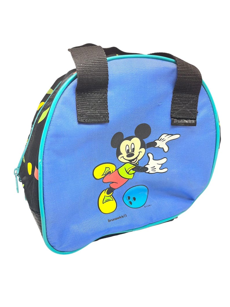 Puede incluir: Una bolsa de bolos azul con un gr&aacute;fico de Mickey Mouse con camisa verde y zapatos amarillos, junto a una bola de bolos azul. La bolsa tiene laterales de tela negra, asas negras y el nombre de la marca Brunswick impreso en blanco en el frente.