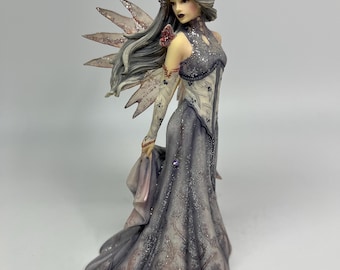 May include: A detailed figurine of a fairy with silver hair and glittery wings. The fairy wears a long, flowing gown in shades of gray and lavender, with intricate detailing. The figure has a serene expression and is holding a piece of fabric.