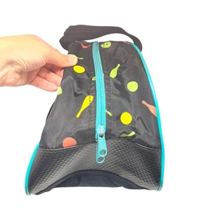 Puede incluir: Una bolsa negra con cremallera que presenta un patr&oacute;n repetitivo de peque&ntilde;os bolos y c&iacute;rculos amarillos, naranjas y verdes. La bolsa tiene un cierre de cremallera turquesa brillante, un asa negra y un panel base negro texturizado.