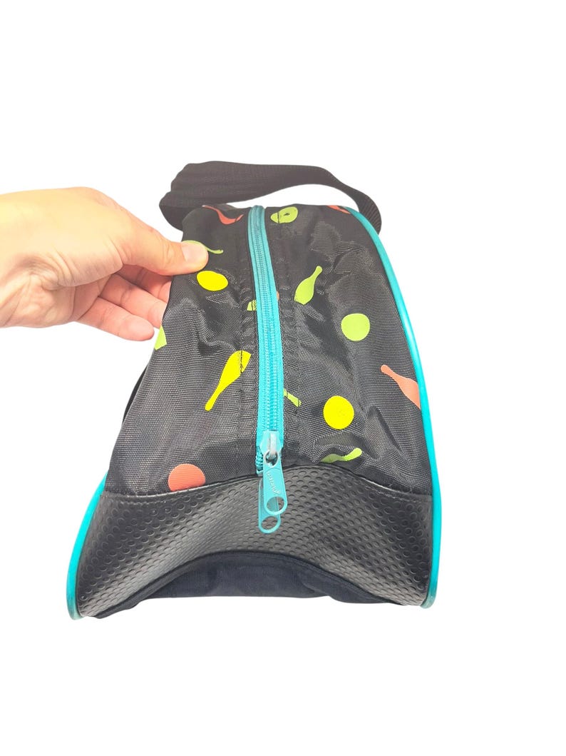 Puede incluir: Una bolsa negra con cremallera que presenta un patr&oacute;n repetitivo de peque&ntilde;os bolos y c&iacute;rculos amarillos, naranjas y verdes. La bolsa tiene un cierre de cremallera turquesa brillante, un asa negra y un panel base negro texturizado.