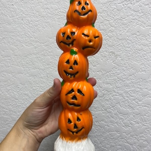 May include: A tall orange candle shaped like a stack of five pumpkins with carved jack-o'-lantern faces. The candle has a white base.