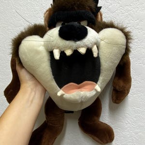 May include: Brown and black plush toy of the cartoon character Taz from Looney Tunes. The toy has a wide open mouth with sharp teeth and a black eye mask.