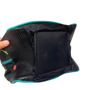 Puede incluir: Un bolso negro con base cuadrada y paneles laterales texturizados. El bolso presenta un ribete en color verde azulado a lo largo de los bordes y un patr&oacute;n abstracto colorido en los laterales. La estructura incluye una base de tela negra s&oacute;lida.