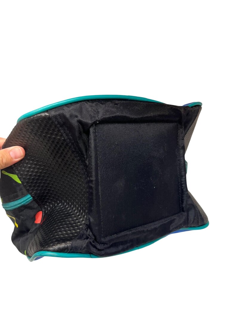 Puede incluir: Un bolso negro con base cuadrada y paneles laterales texturizados. El bolso presenta un ribete en color verde azulado a lo largo de los bordes y un patr&oacute;n abstracto colorido en los laterales. La estructura incluye una base de tela negra s&oacute;lida.