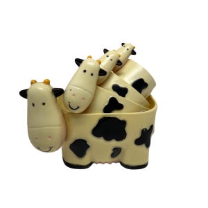 May include: A set of cream-colored measuring cups shaped like a cow. The cups have black spots and cow head handles. The set includes four cups of varying sizes, stacked together. The cow's face has a pink nose and a smiling mouth.