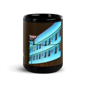Puede incluir: Una taza de cerámica negra con una impresión fotográfica de un edificio de varias plantas iluminado con una luz azul brillante por la noche. La estructura tiene una fachada curva con múltiples ventanas rectangulares y una plataforma.
