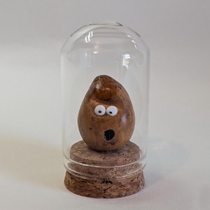 May include: A brown, egg-shaped object with a smooth texture, featuring two small white googly eyes and a dark, circular open mouth. The object is positioned on a circular cork base and is enclosed within a clear, cylindrical glass dome.