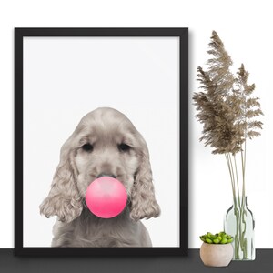 Op de afbeelding: Een ingelijste print van een lichtbruine hond met roze kauwgom in zijn bek. De hond kijkt recht in de camera.
