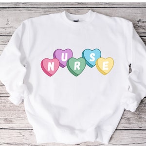 White crewneck sweater lying flat on the floor. Sweater has 5 candy hearts with the letters stating "NURSE"