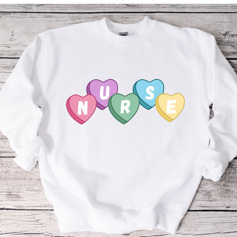 White crewneck sweater lying flat on the floor. Sweater has 5 candy hearts with the letters stating "NURSE"