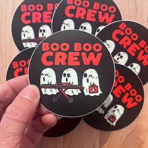 Könnte beinhalten: Ein Satz von sechs runden Aufklebern mit schwarzem Hintergrund und weißen Geisterillustrationen. Die Aufkleber zeigen den Text "BOO BOO CREW" in roten Buchstaben.