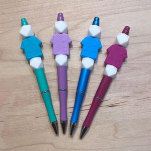 Puede incluir: Cuatro bolígrafos coloridos con un diseño de bata médica. Los bolígrafos son de color turquesa, morado, azul y rosa.