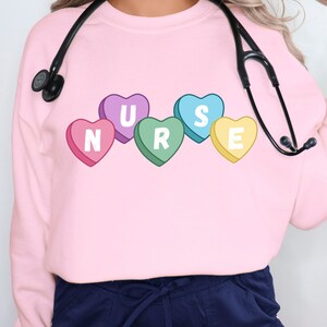 Female nurse wearing a pink crewneck sweater. Sweater has 5 candy hearts with the letters stating "NURSE"