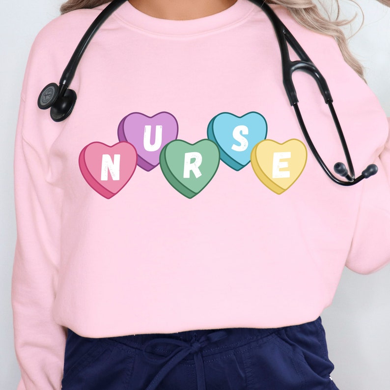 Female nurse wearing a pink crewneck sweater. Sweater has 5 candy hearts with the letters stating "NURSE"