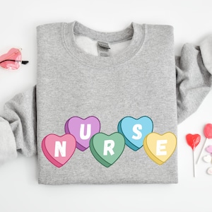 Sport Grey crewneck  sweater folded lying on floor. Sweater has 5 candy hearts with the letters stating "NURSE"