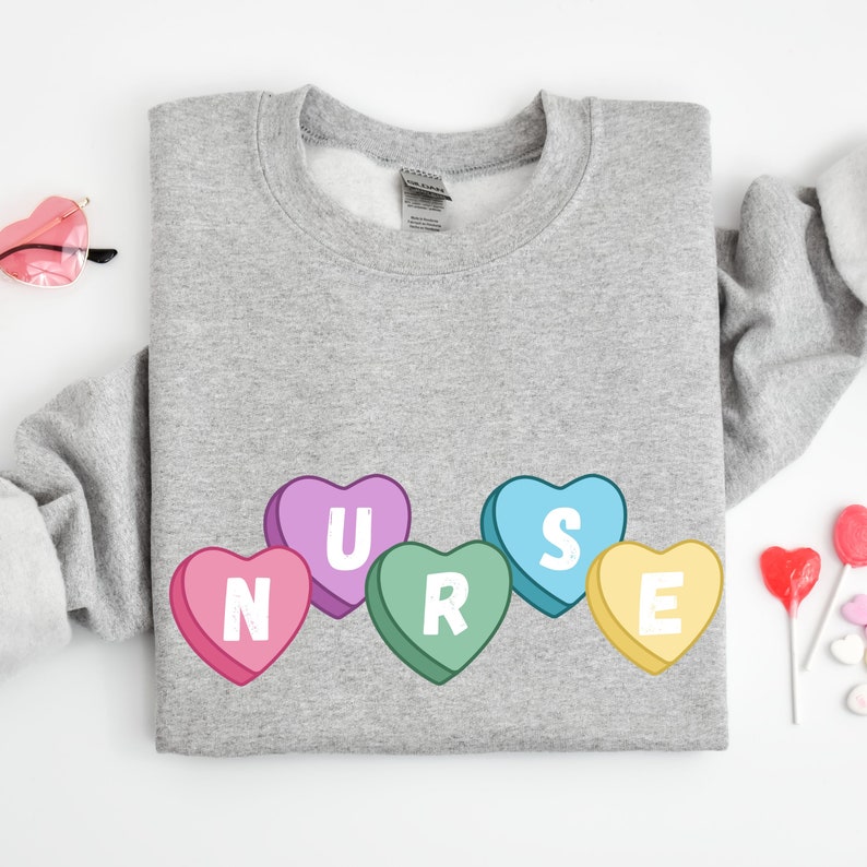 Sport Grey crewneck  sweater folded lying on floor. Sweater has 5 candy hearts with the letters stating "NURSE"