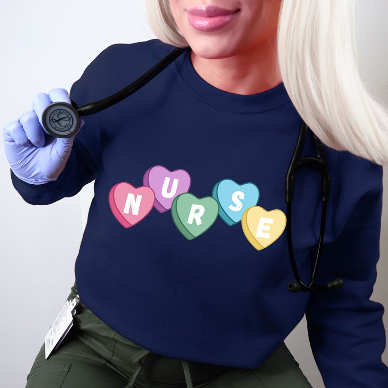 Female nurse wearing a navy blue crewneck sweater. Sweater has 5 candy hearts with the letters stating "NURSE"