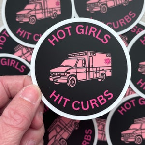 Könnte beinhalten: Ein runder Aufkleber mit schwarzem Hintergrund und einer rosa Krankenwagen-Illustration. Der Text "HOT GIRLS HIT CURBS" ist in Rosa am Rand des Aufklebers. Mehrere Aufkleber sind sichtbar.
